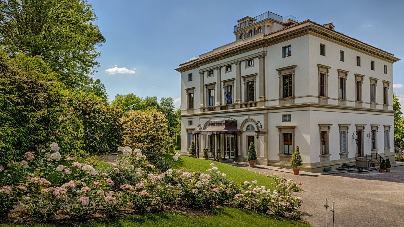 Elegant villa with cream walls, large windows, and a porch. Lush garden with blooming pink and white flowers under a clear blue sky.