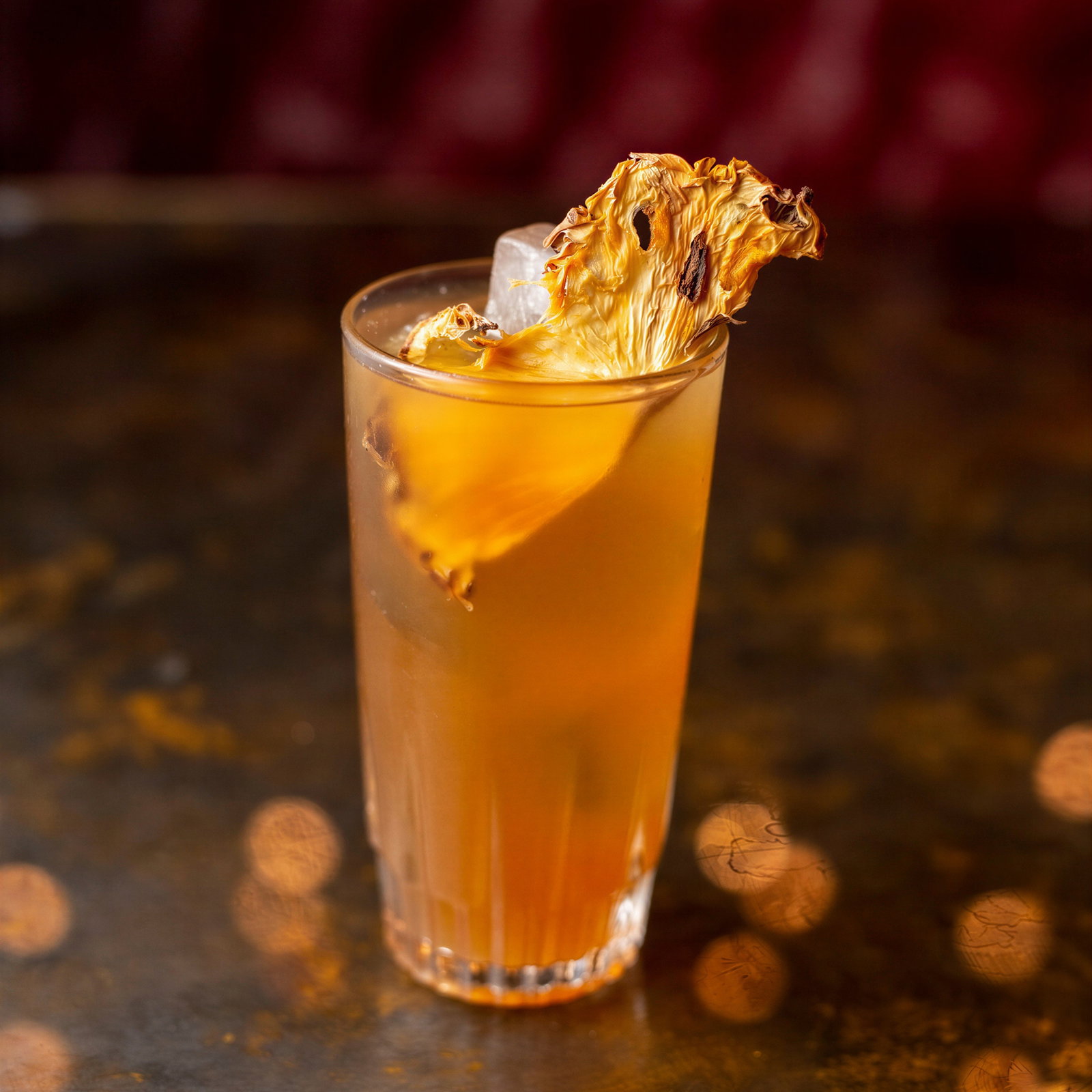 A tall glass of amber-colored drink with ice and a dried pineapple garnish. The setting has a warm, dimly lit ambiance with bokeh lights.