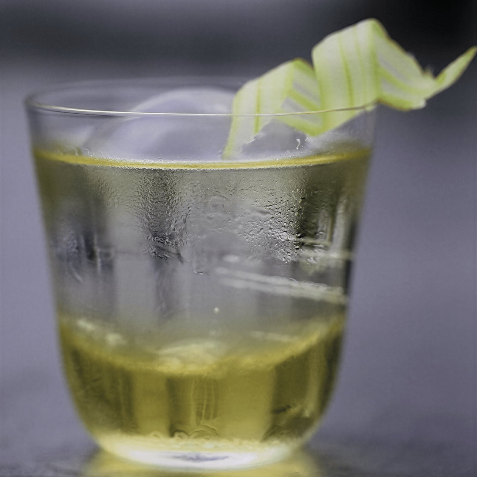 A glass of yellow drink with a large ice cube and a green ribbon garnish on a smooth surface, set against a blurred background.