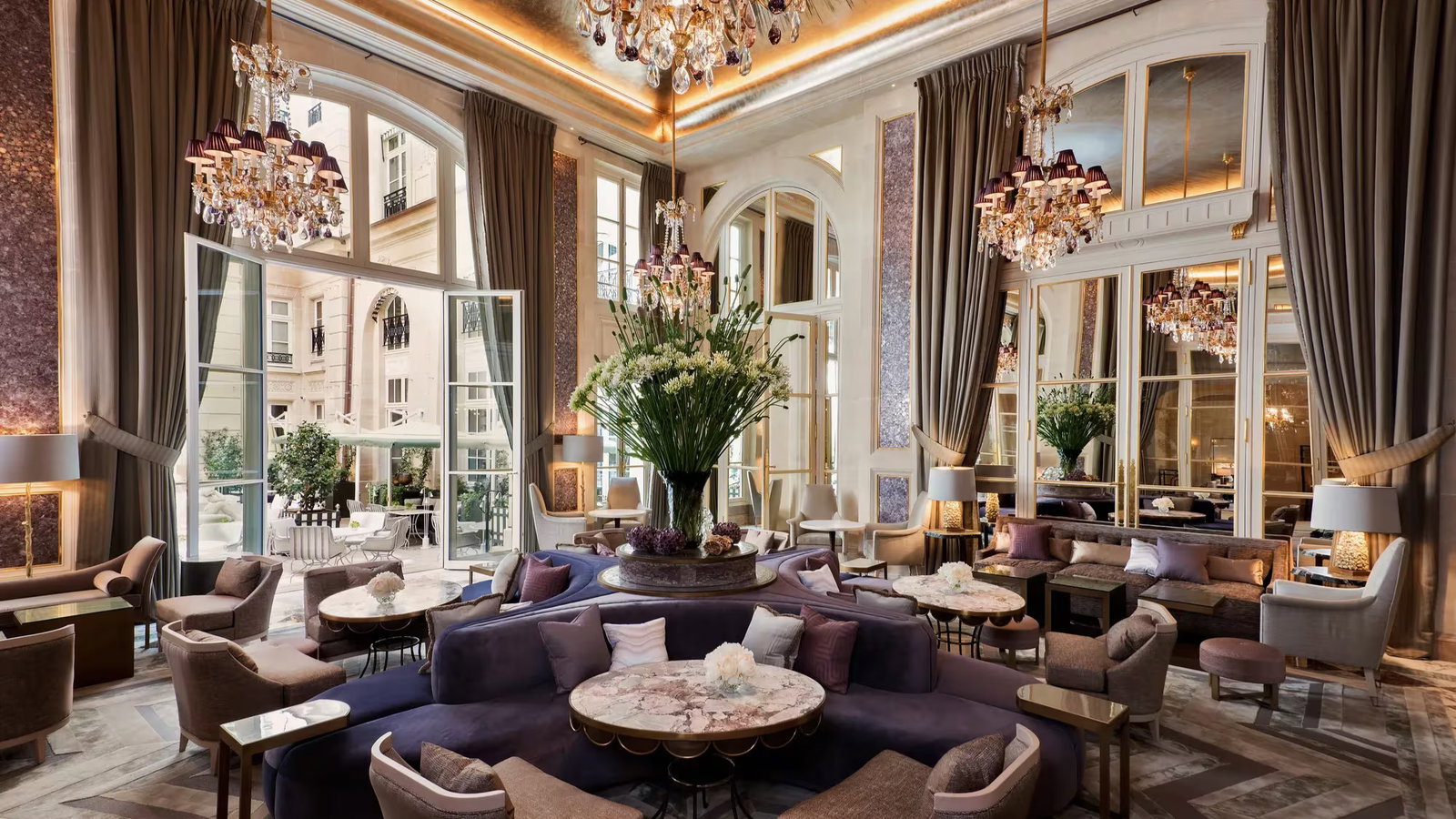 Elegant hotel lounge with plush seating in purple and beige tones, large floral centerpiece, chandeliers, and open patio doors. Luxurious ambiance.