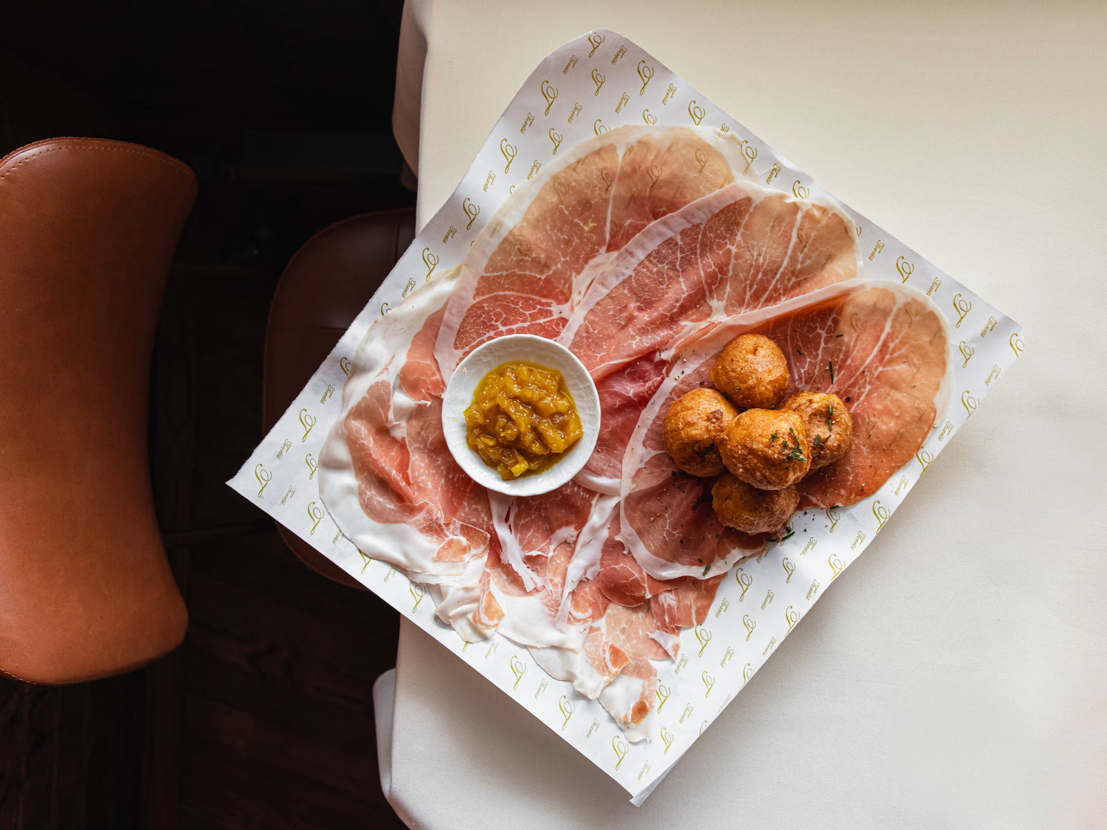 Prosciutto slices and fried snacks on patterned paper with a bowl of relish on a table with brown chairs. Light and elegant setting.