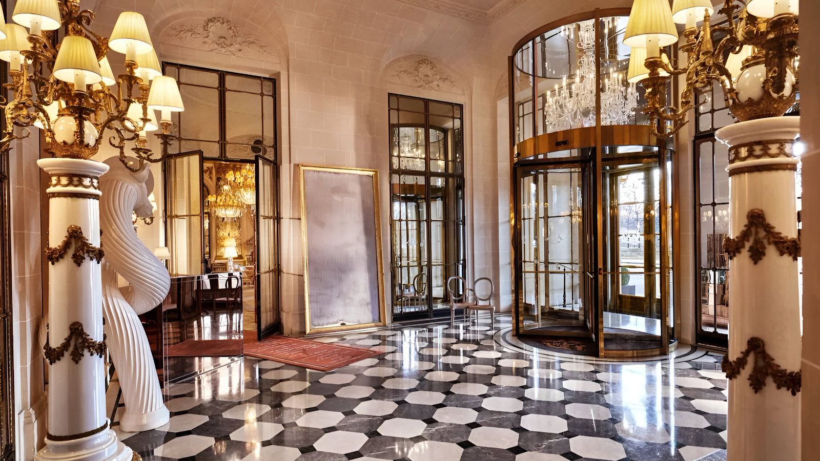 Elegant lobby with a revolving door, chandelier, marble columns, and checkered floor. Art piece leaning on wall, warm lighting.