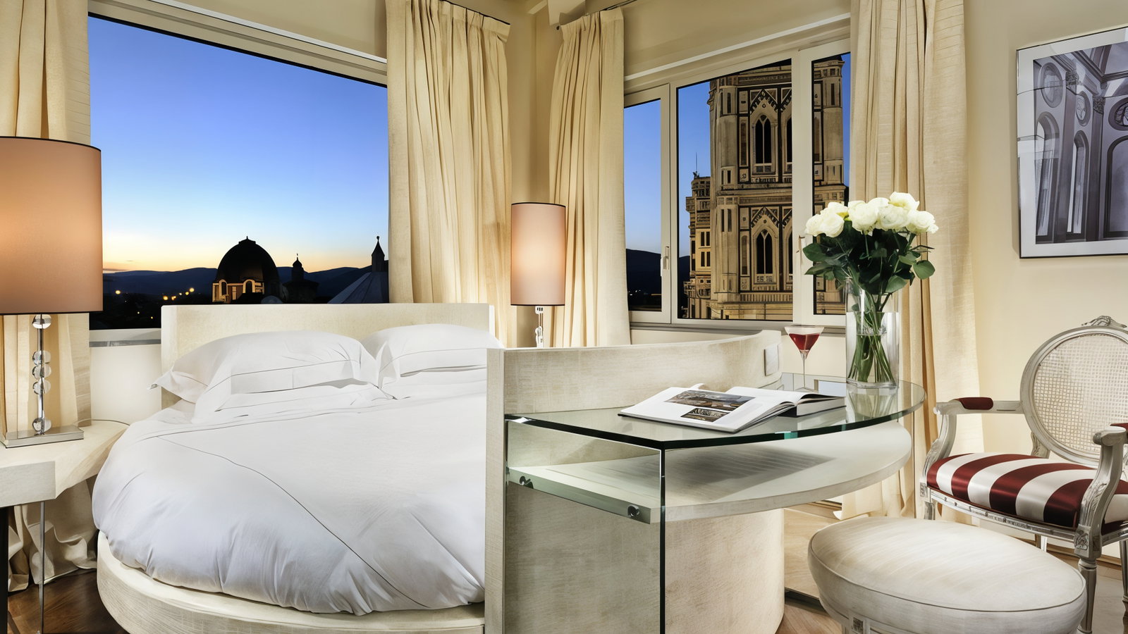 Elegant bedroom with a round bed, white linens, glass table, roses in a vase, and a view of a sunset cityscape through large windows.