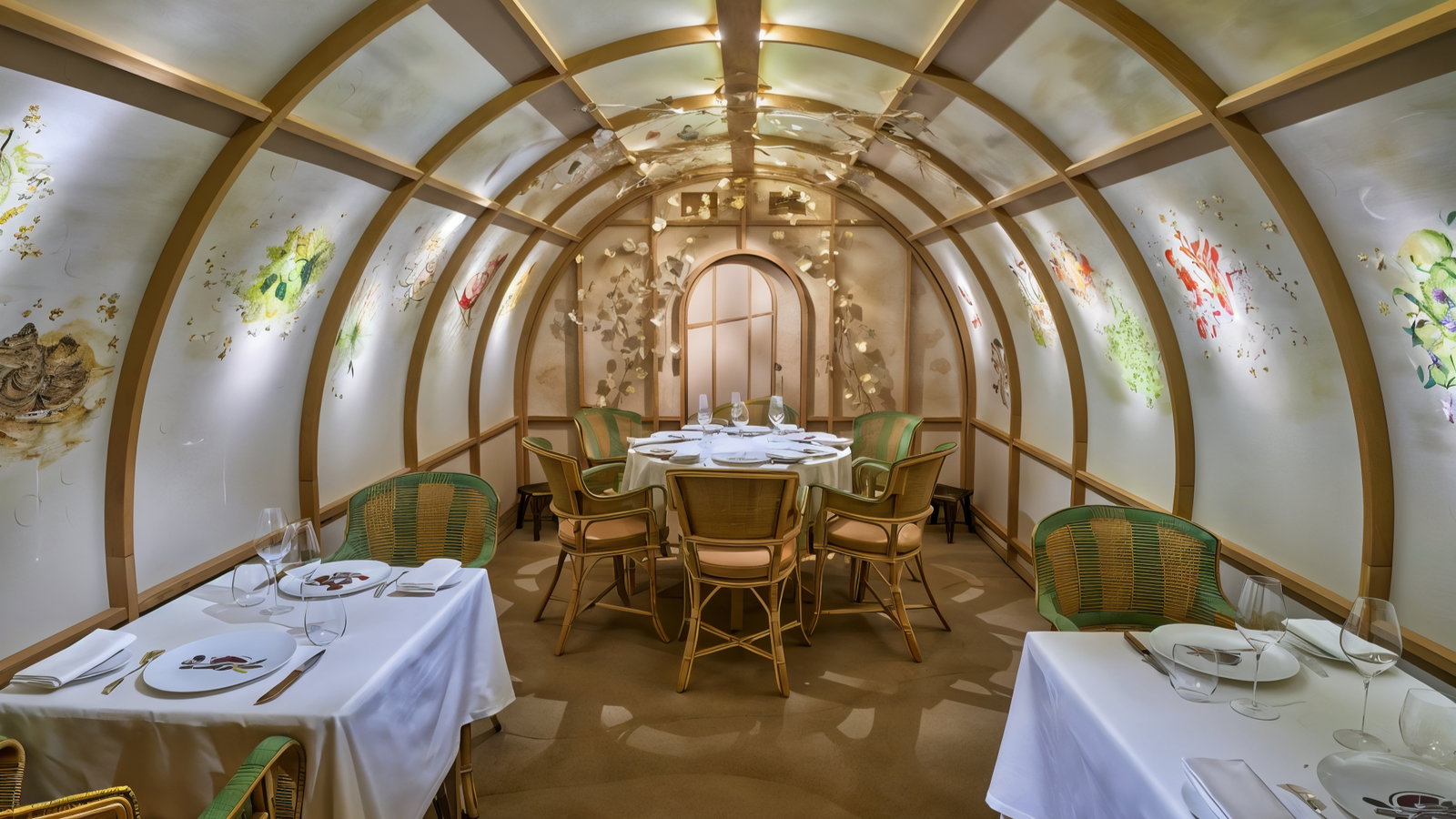 Elegant restaurant with arched walls adorned with floral art. Round table set for dining; green chairs, warm lighting, cozy ambiance.