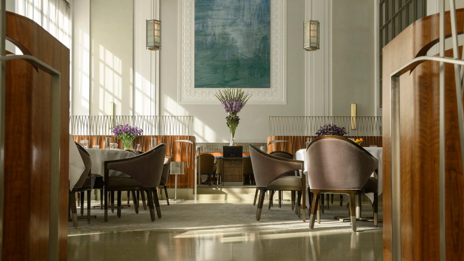 Elegant restaurant with round tables, brown chairs, and floral centerpieces. Large blue artwork and tall windows create a serene ambiance.