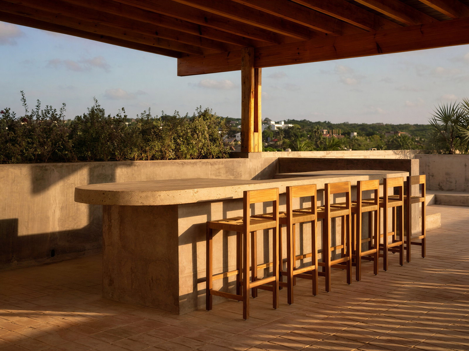 Outdoor Rooftop Sunset bar at Hotel Humano with wooden stools under a wooden pergola. Sunlight casts long shadows on the tiled floor. Greenery and sky in the background.