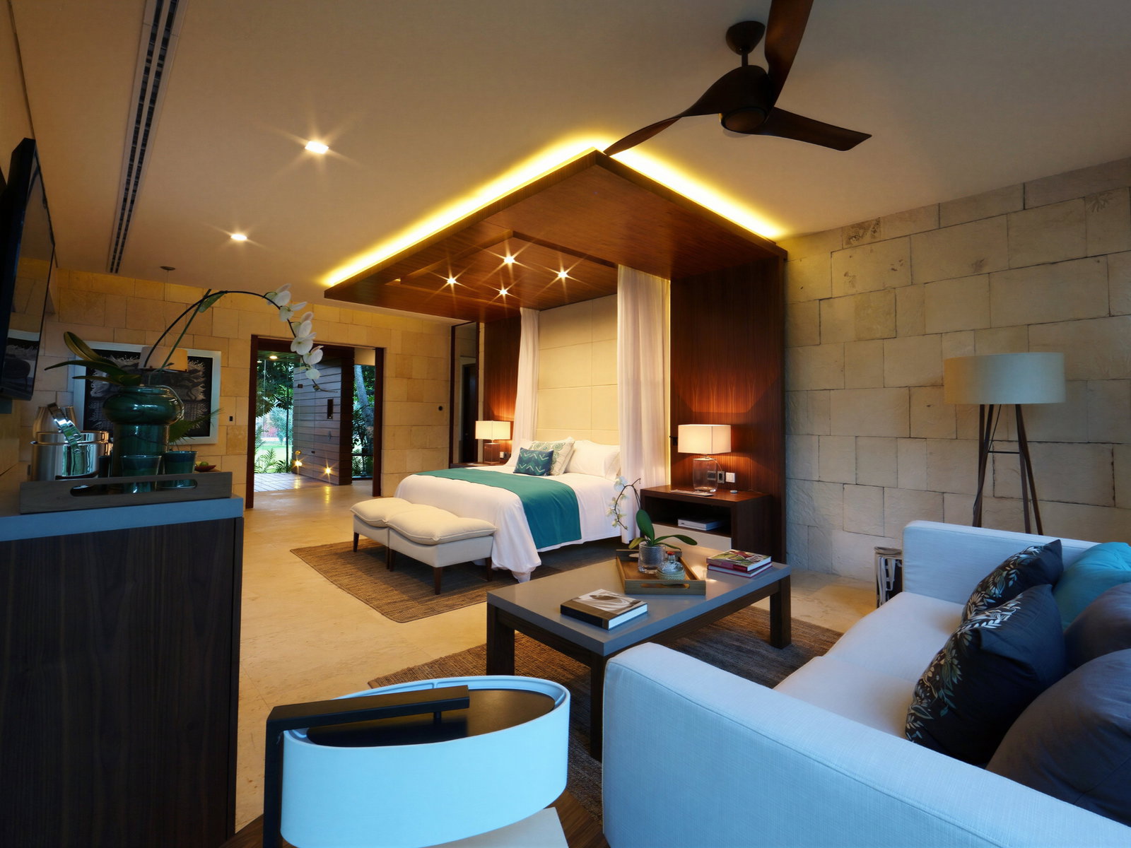 Luxurious bedroom with a canopy bed, teal accents, and wooden decor. Soft lighting sets a cozy mood. White orchids and books adorn tables.