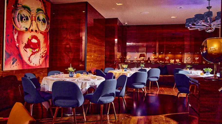 Modern restaurant with dark wood walls, blue chairs, and white tablecloths. Vibrant mural of a woman in glasses on the wall, warm lighting.