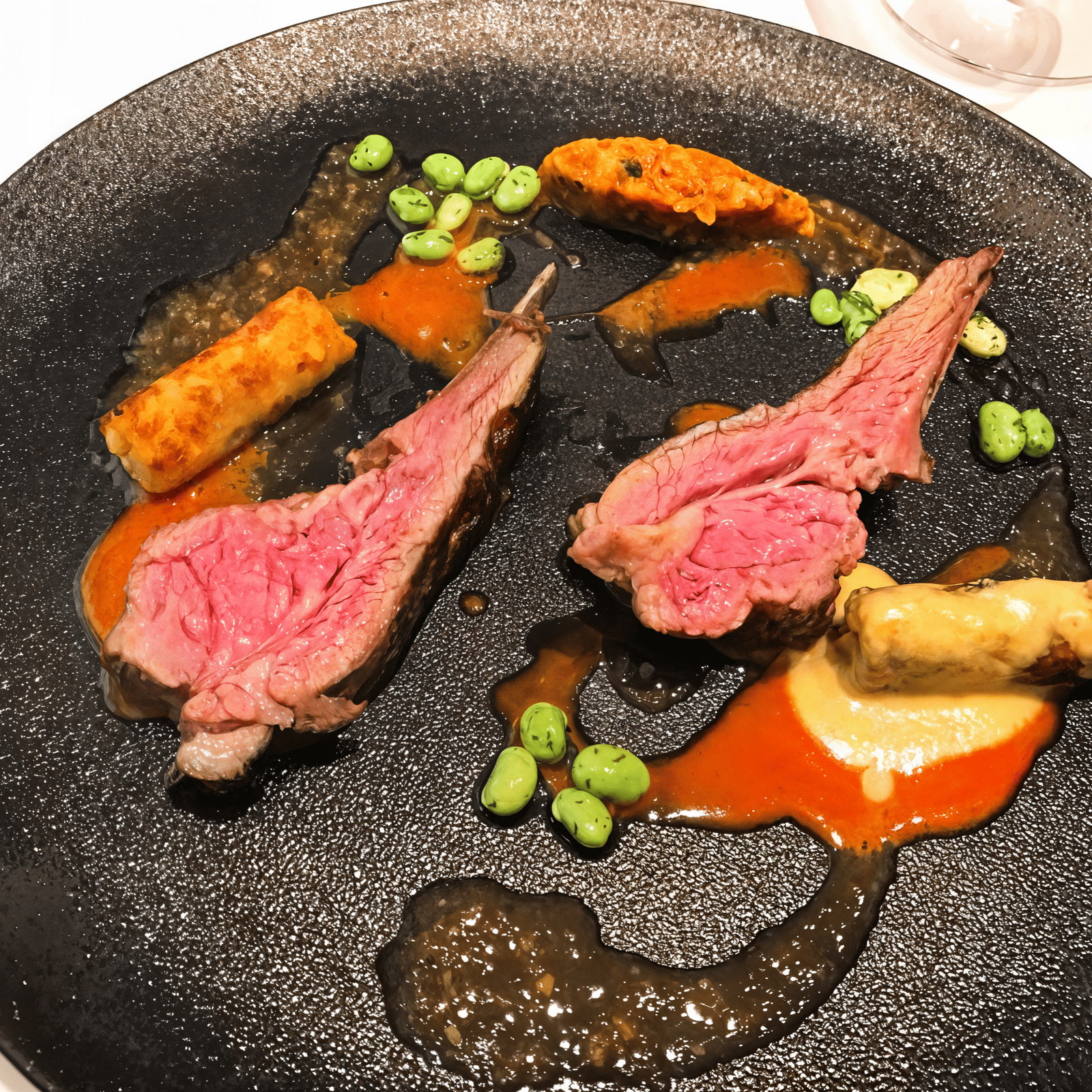 Gourmet dish with two lamb chops, green edamame, crispy rolls, and vibrant sauces on a dark textured plate. Elegant and appetizing.