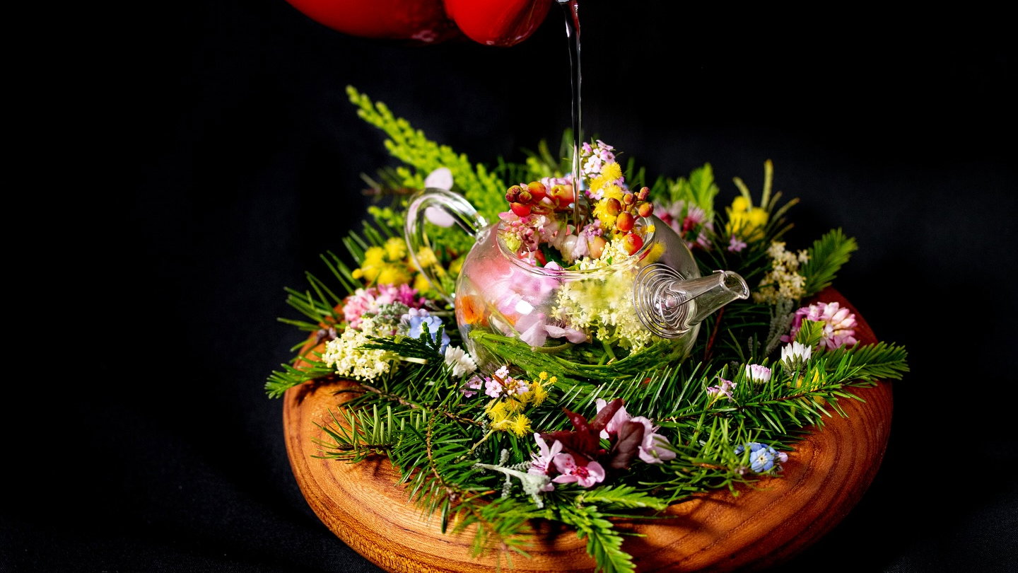 Transparent teapot filled with colorful flowers and greenery on wooden board. Red liquid being poured in. Dark background enhances hues.