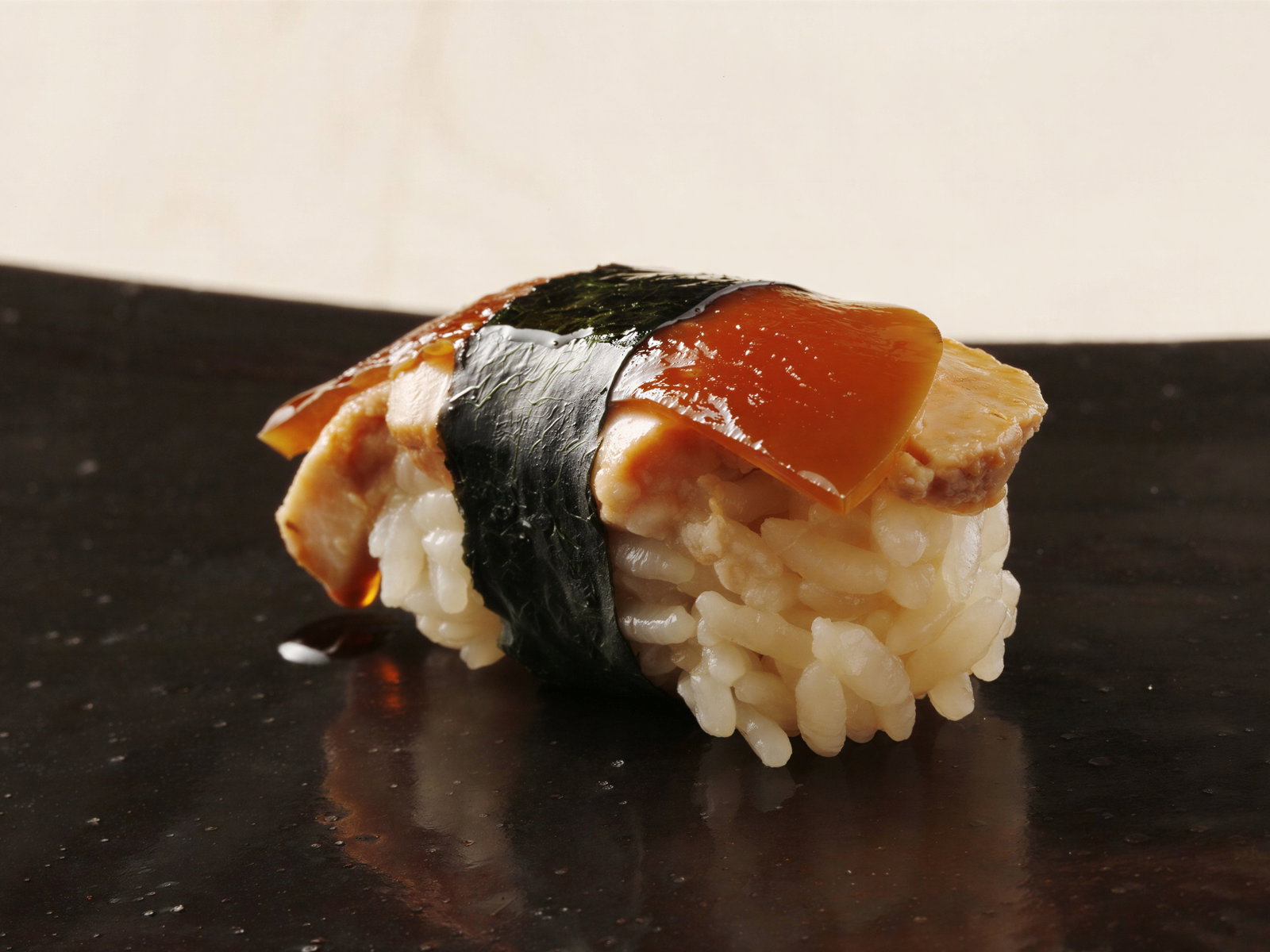 Close-up of a sushi piece with rice, amber topping, and nori, on a dark, glossy plate. Minimalist and elegant display.