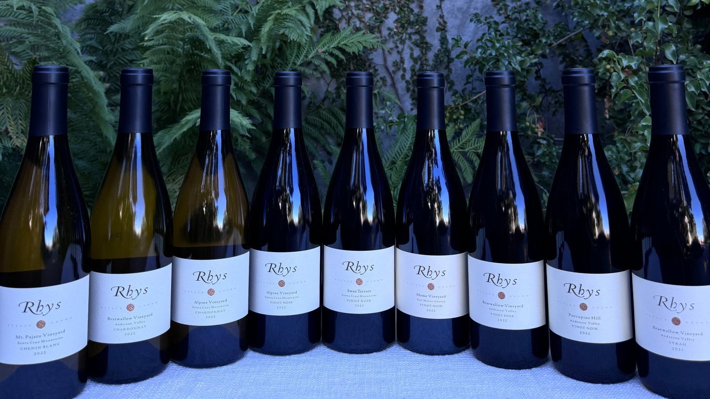 A row of Rhys wine bottles outdoors, displayed on a white cloth. Green foliage in the background. Labels indicate various vineyards and years.