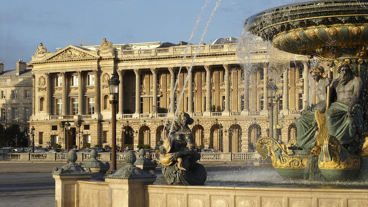 Fountain with ornate statues spraying water, set against a grand historic building under a clear blue sky, evoking elegance and grandeur.