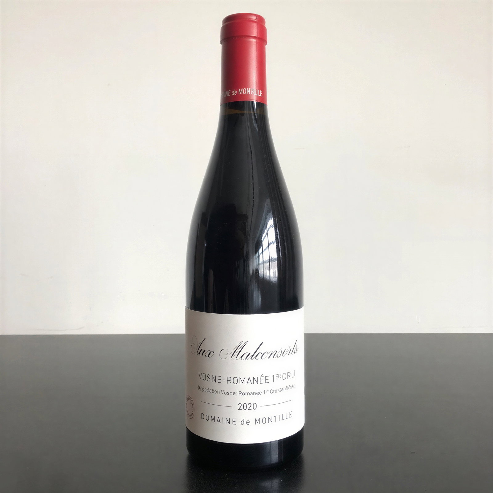 A bottle of Domaine de Montille Aux Malconsorts 2020 wine, with a red cap, stands on a dark surface against a plain background.