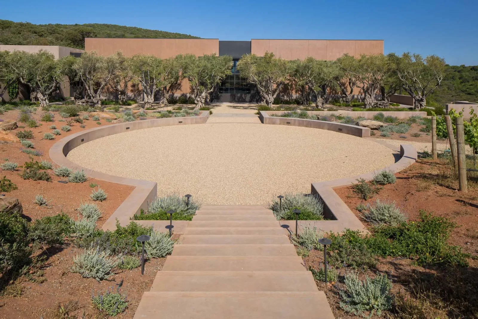 Stone steps lead to a circular gravel courtyard, surrounded by greenery and olive trees. Modern building facade in the background under blue sky.