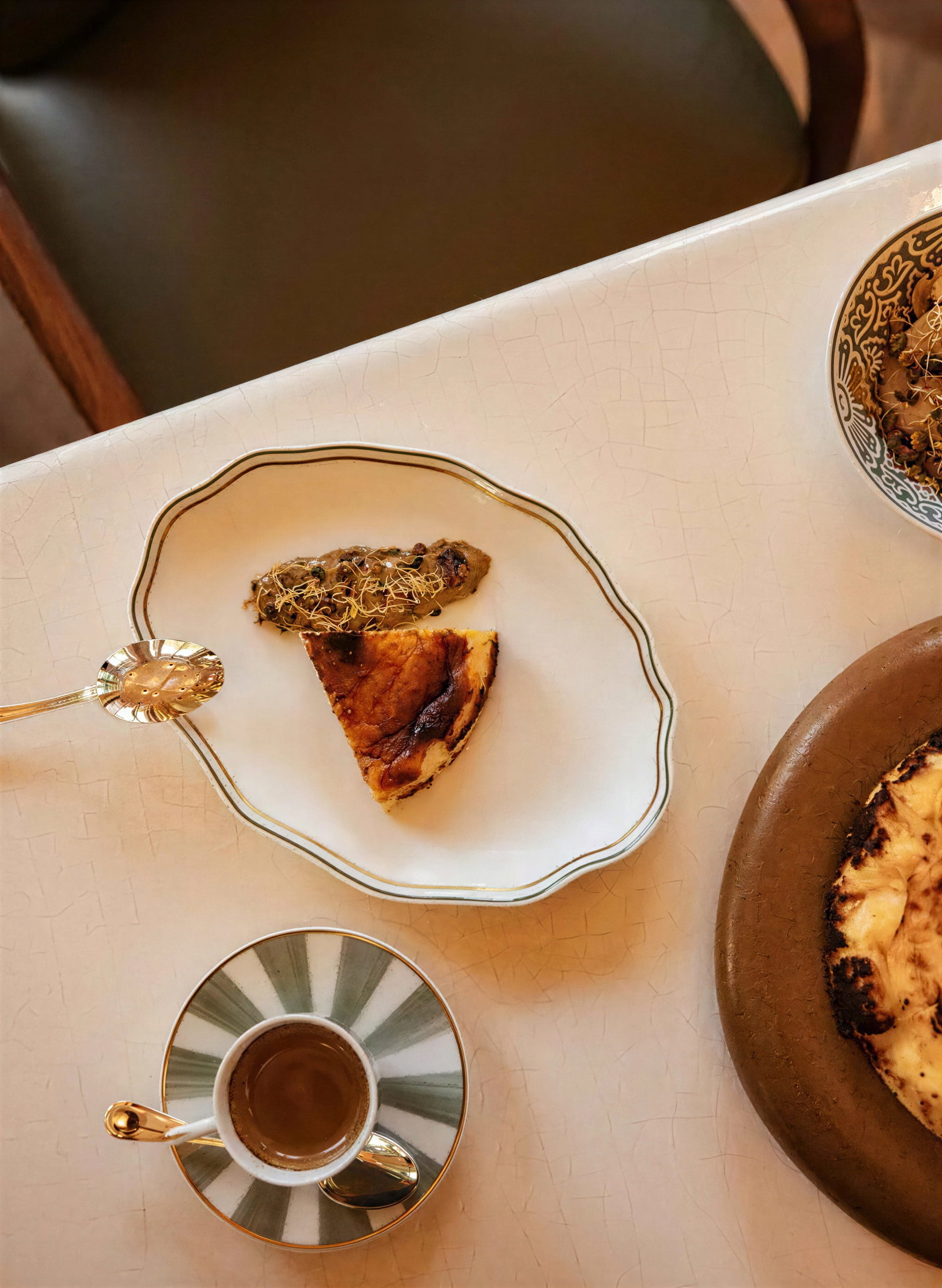 Slice of pistachio honey cheesecake from Serra at The Chancery Rosewood and topping on a decorative plate, next to a green-striped cup of espresso on a white table. Cozy, inviting atmosphere.