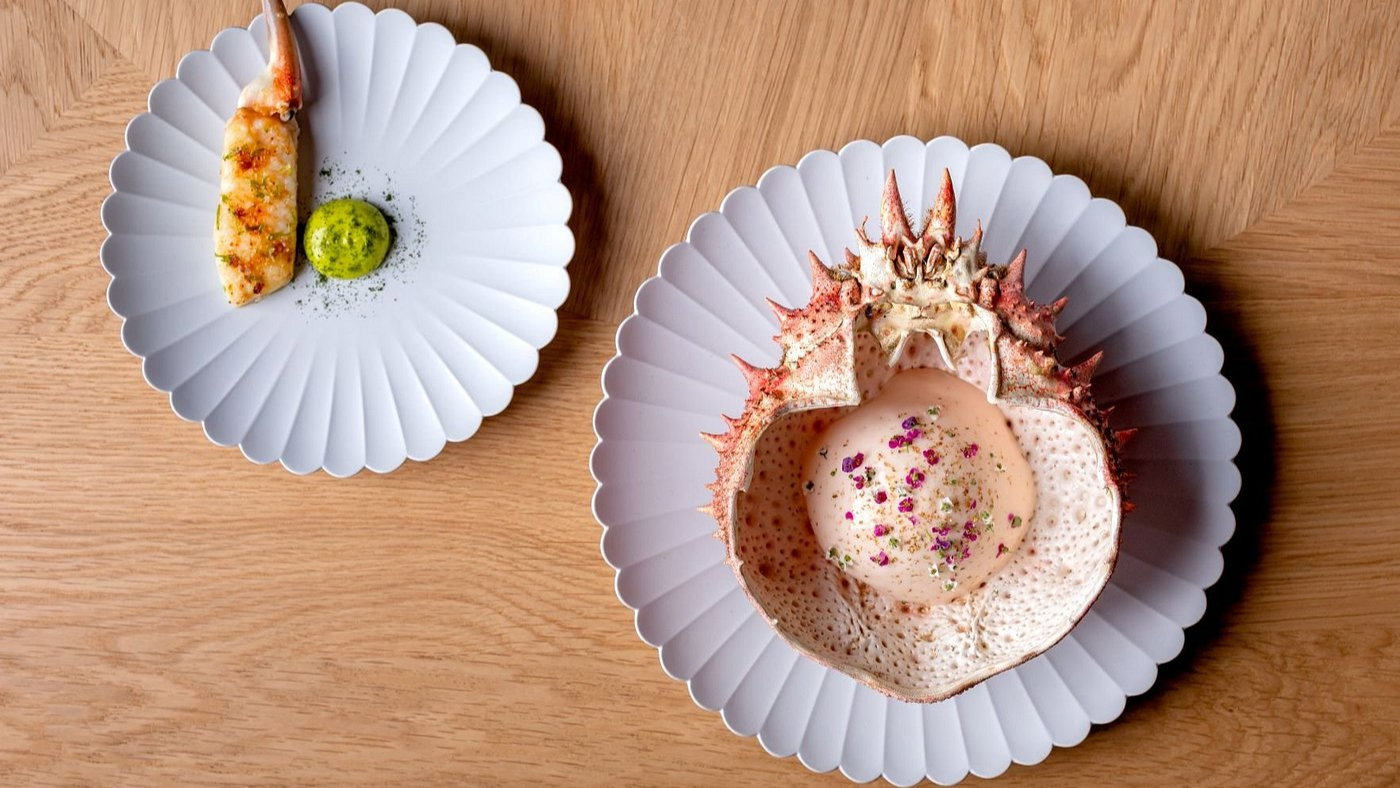 Crab shell with creamy dish and herbs on a fluted white plate, next to a crab leg and green sauce on another plate, on a wooden table.