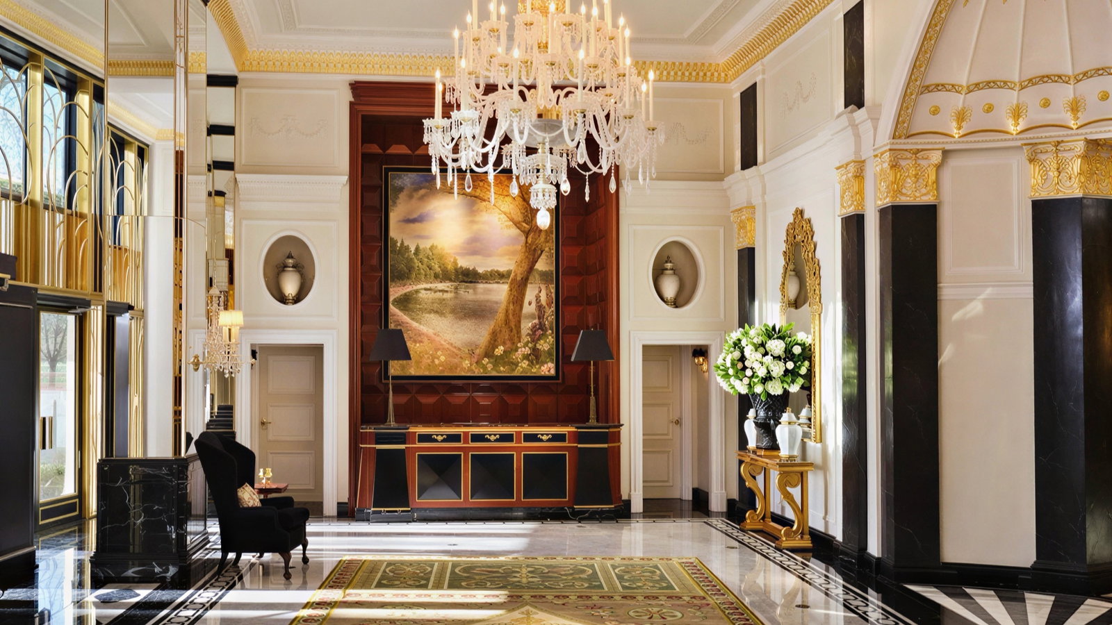 Elegant hotel lobby with a large chandelier, a painting of a lake, black marble columns, gold accents, and a black chair on a patterned rug.