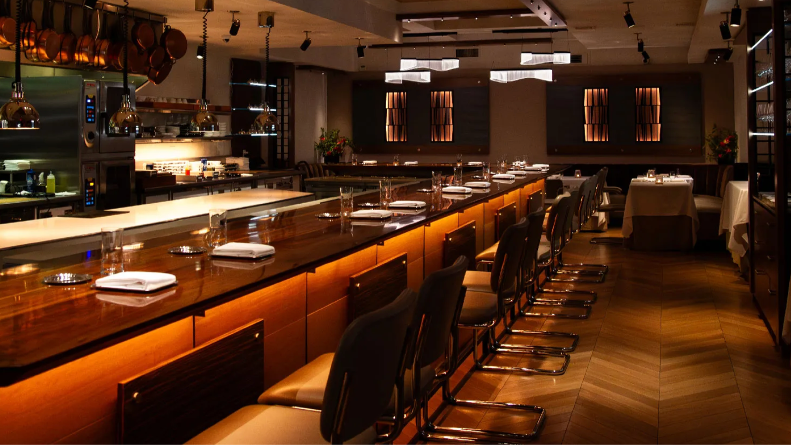 Cozy restaurant interior with a long wooden bar, empty chairs, and dim lighting. Copper pans hang in the kitchen; elegant dining setup.