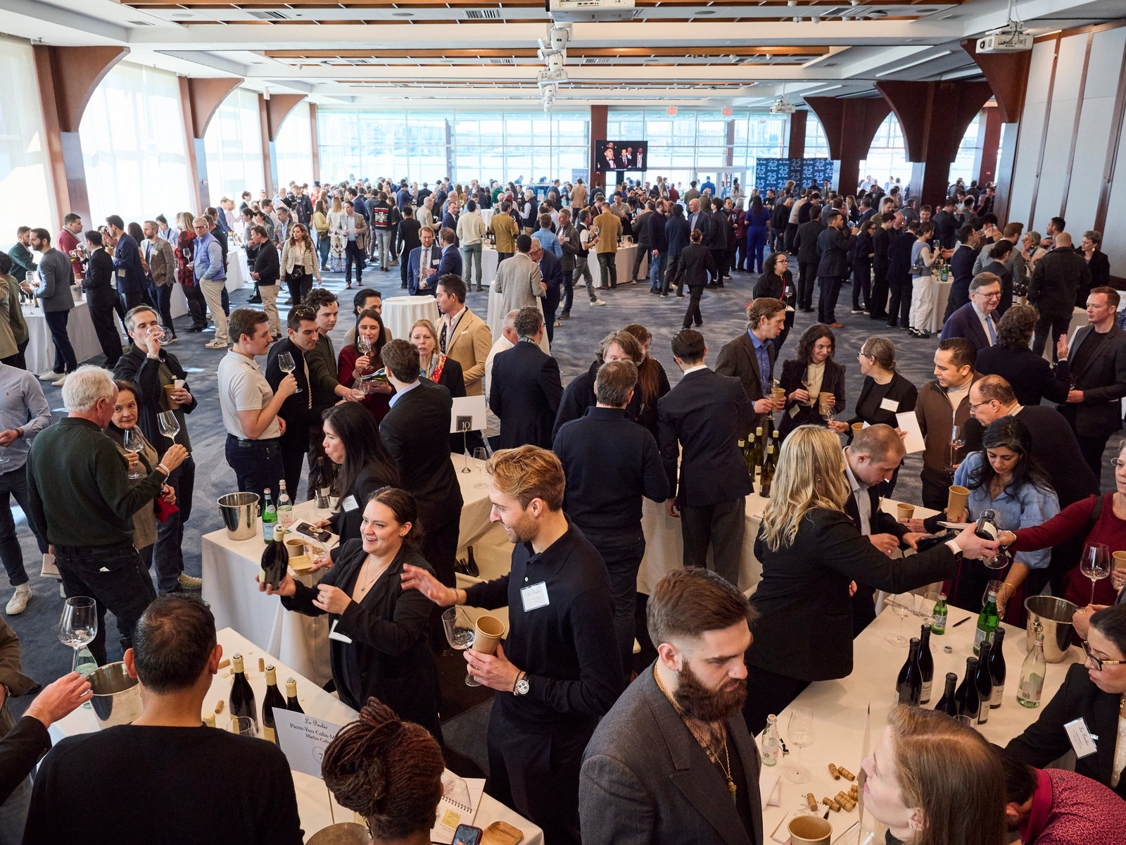 Bright Grand Tasting room at La Paulée NYC 2025. Guests socialize, sampling wine at tables. Attendees wear name badges, creating a lively atmosphere.