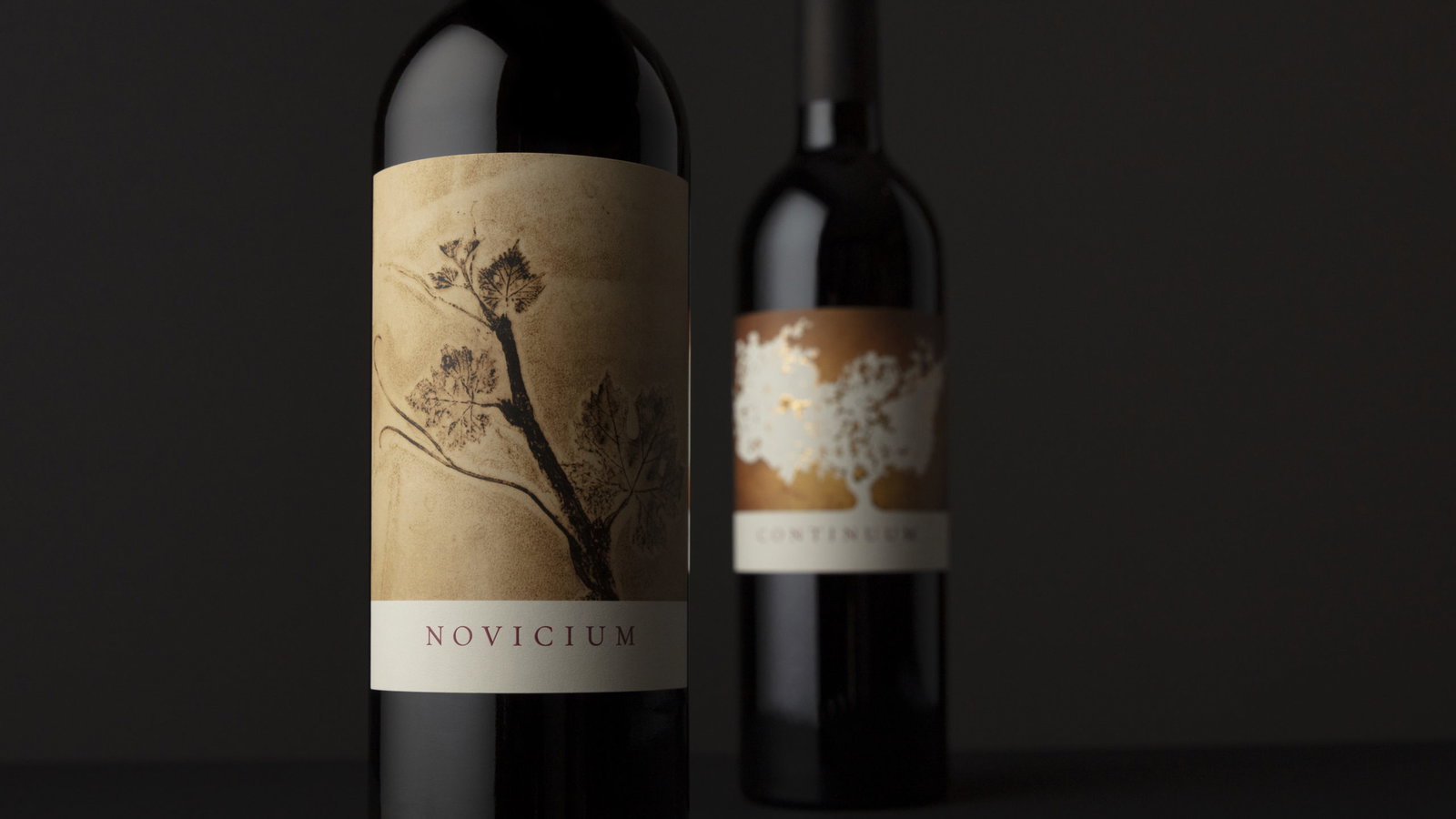 Two wine bottles with elegant labels on a dark background. The front label shows a branch, text "NOVICIUM." The rear label has a tree.