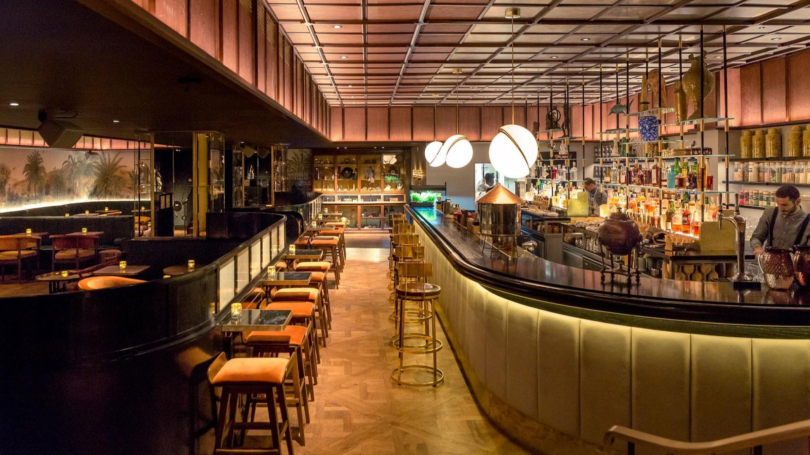 Stylish bar interior with dim lighting, wooden decor, and globe lights. Bartenders behind the counter; empty stools and tables set mood.
