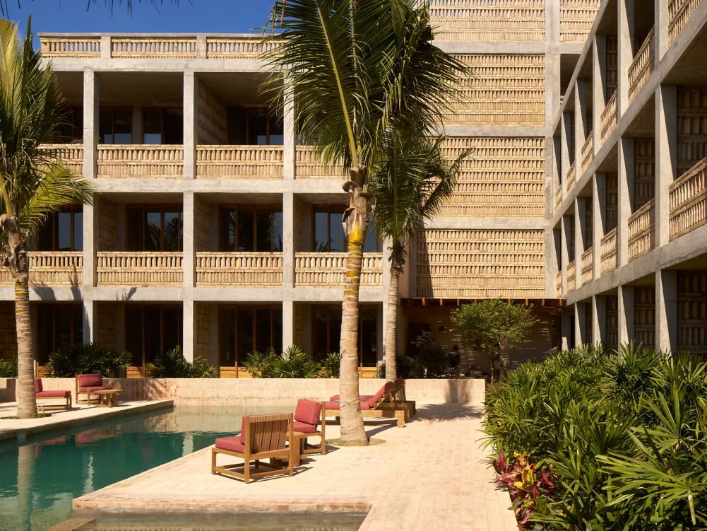 The sunlit tropical pool courtyard of Hotel Humano with palm trees, a turquoise pool, and red-cushioned wooden chairs. Beige geometric building in background.