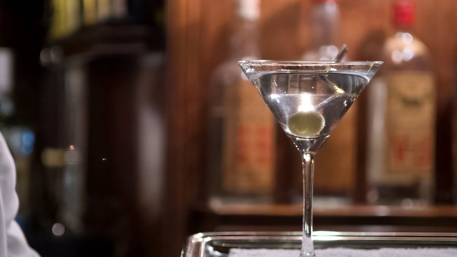 A waiter holds a tray with a martini glass and an olive in a dimly lit bar with blurred liquor bottles in the wooden background.