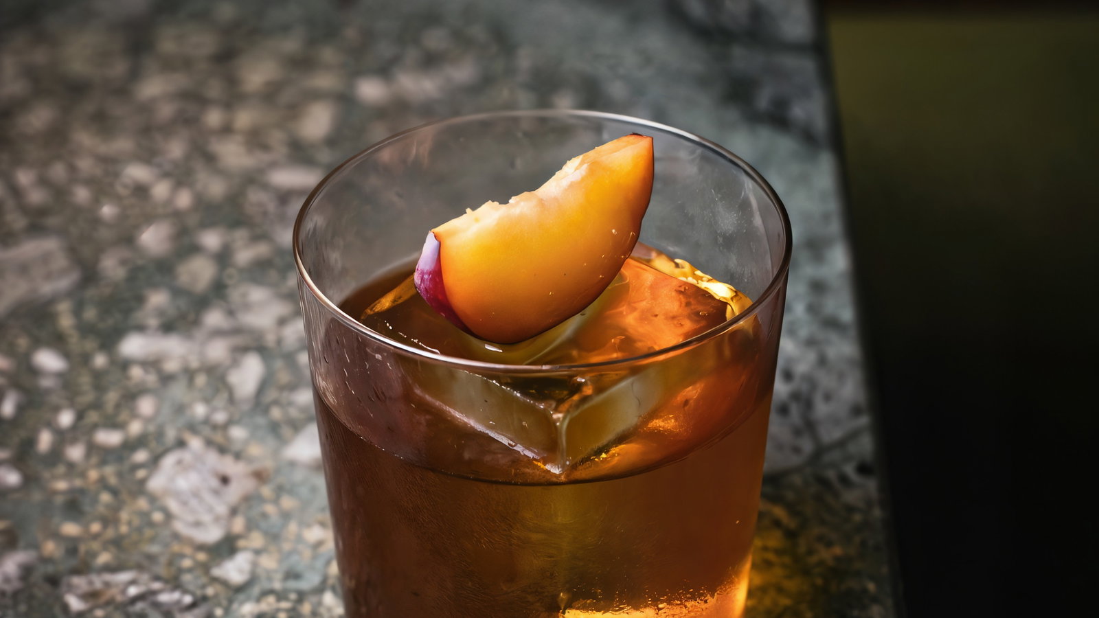 A glass with amber liquid and ice, topped with a peach slice, on a textured gray stone surface. The mood is elegant and serene.