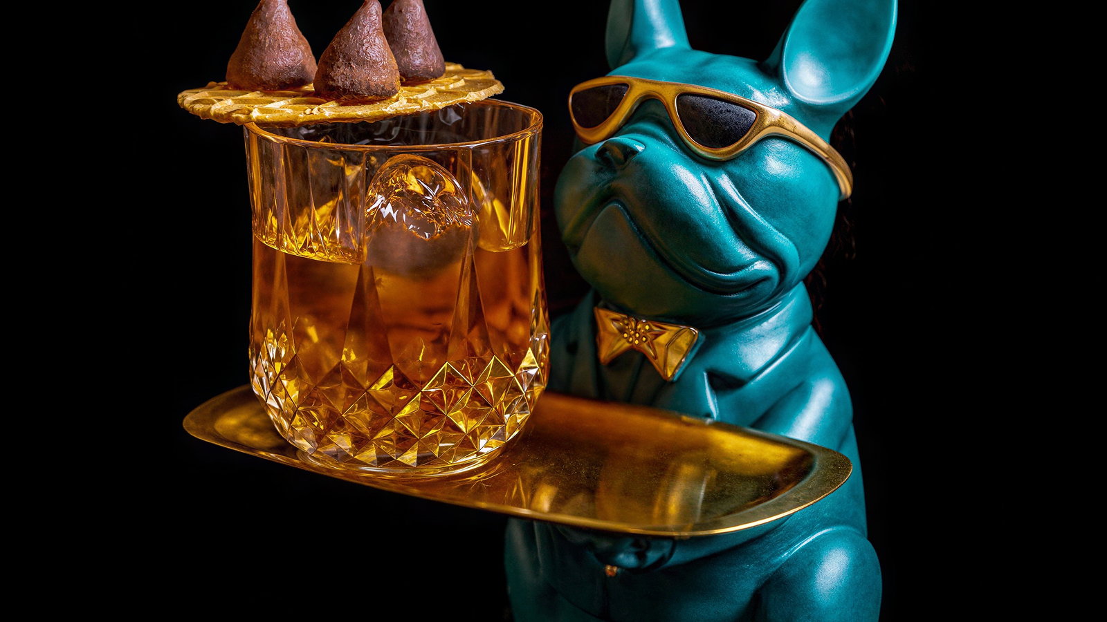 Turquoise bulldog figurine wearing sunglasses holds gold tray with a whiskey glass and waffle topped with chocolates on a black background.