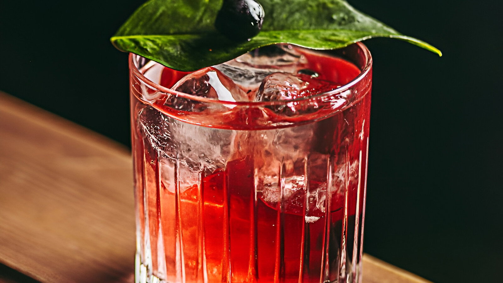 Red cocktail in a glass with ice, garnished with a green leaf and black berry, sits on a wooden bar against a dark background.