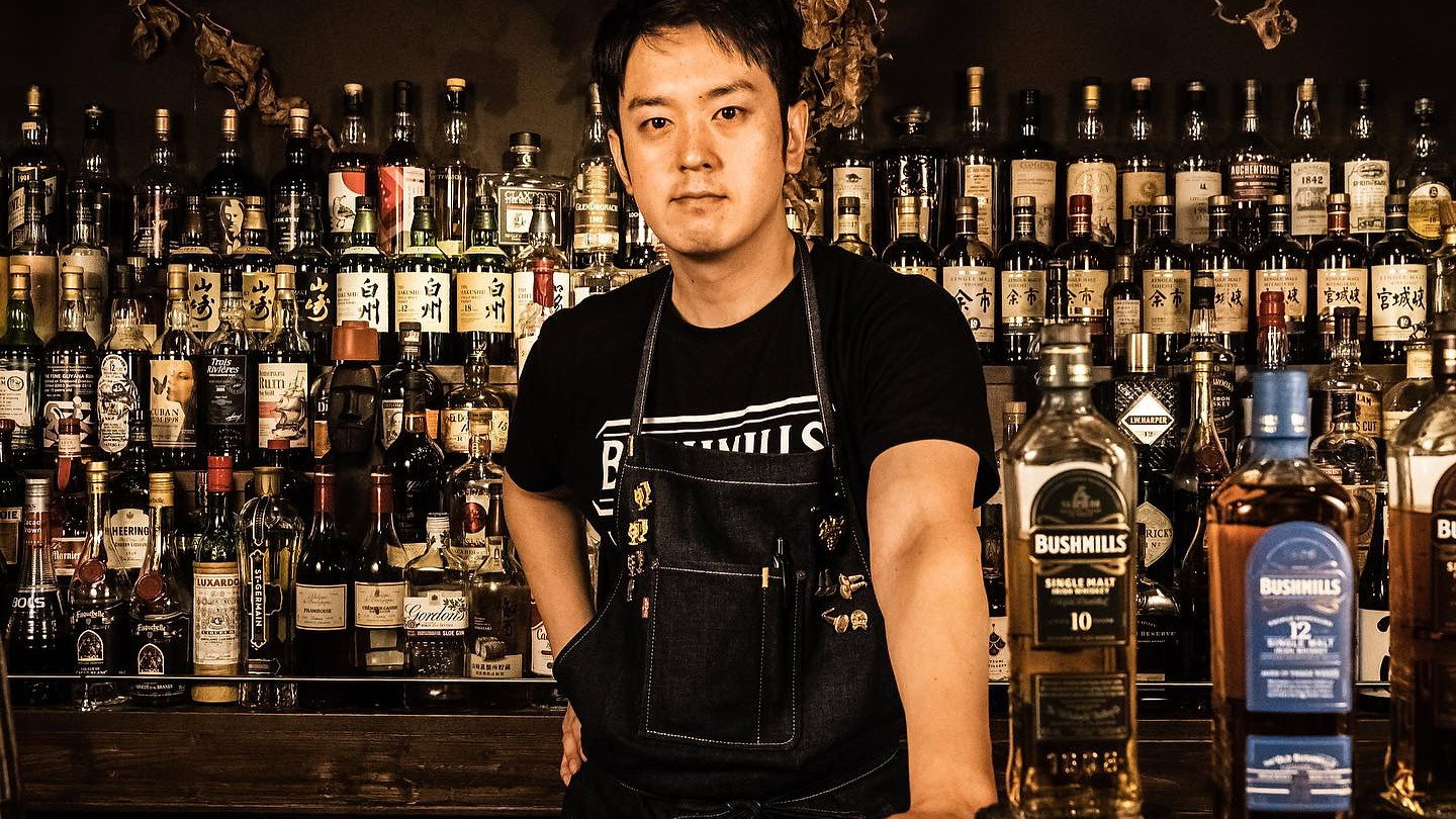 Bartender in a dark bar, wearing a denim apron, stands confidently. Shelves of liquor bottles in background; Bushmills bottles on counter.