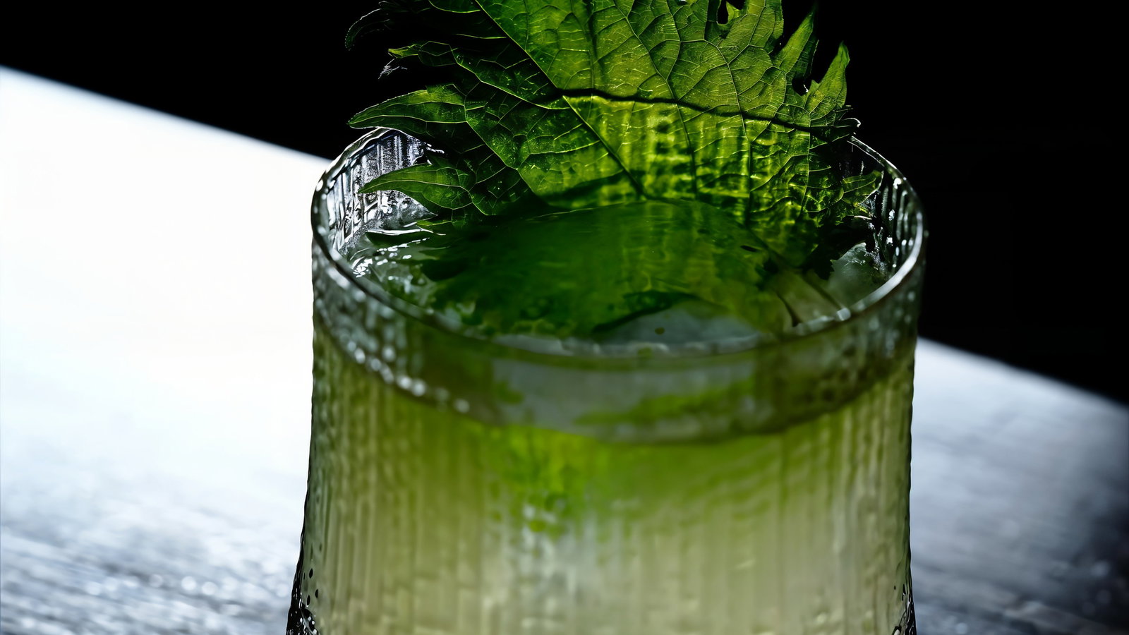 Textured glass of light green liquid with ice, topped with vibrant green leaves, on a dark wooden table, under dramatic lighting.