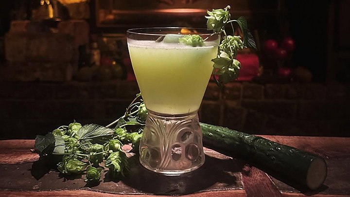 A green cocktail in a decorative glass, garnished with hops, sits on a wooden bar. A cucumber lies beside it, with a dim, cozy background.