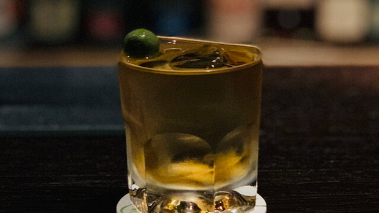 A glass of whiskey with ice and an olive on a coaster, set on a dark bar counter against a blurred background of bottles, dimly lit.