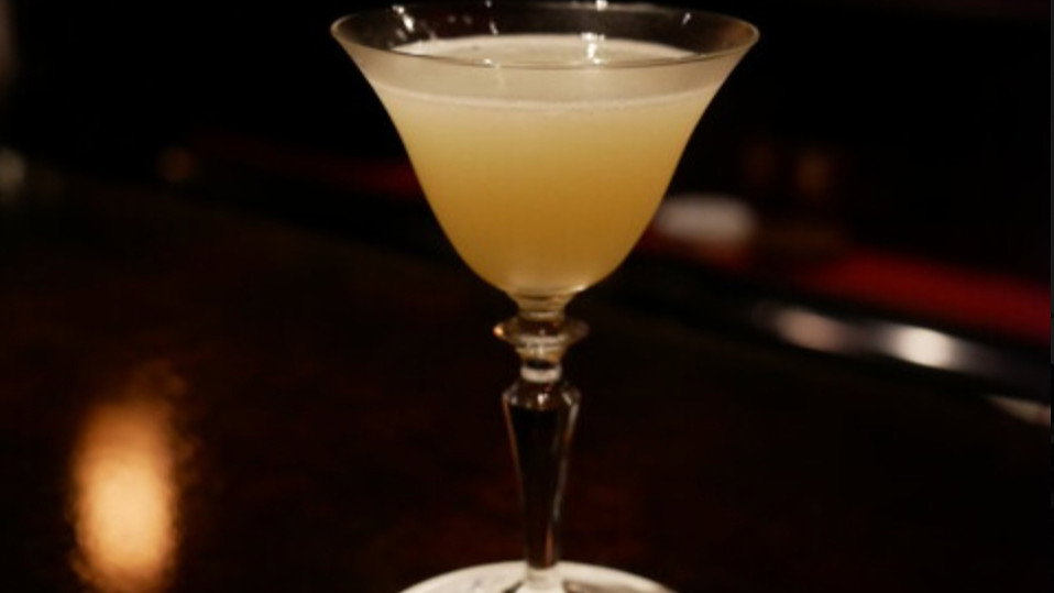 A lemon-yellow cocktail in a martini glass on a dark bar counter, set on a white coaster. Dim and cozy atmosphere.
