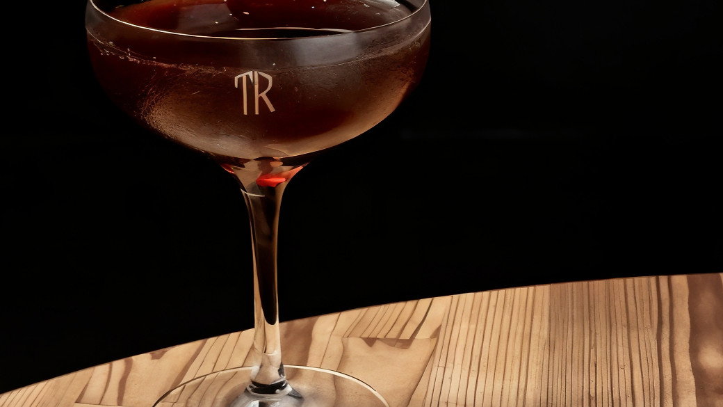 A dark brown cocktail in a glass with "TR" logo, placed on a wooden table. Black background enhances its elegant presentation.