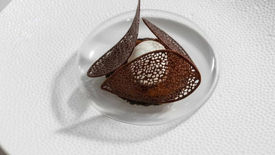 Elegant dessert with intricate chocolate lace decor atop a scoop of cream, served on a textured white plate. Minimalist presentation.