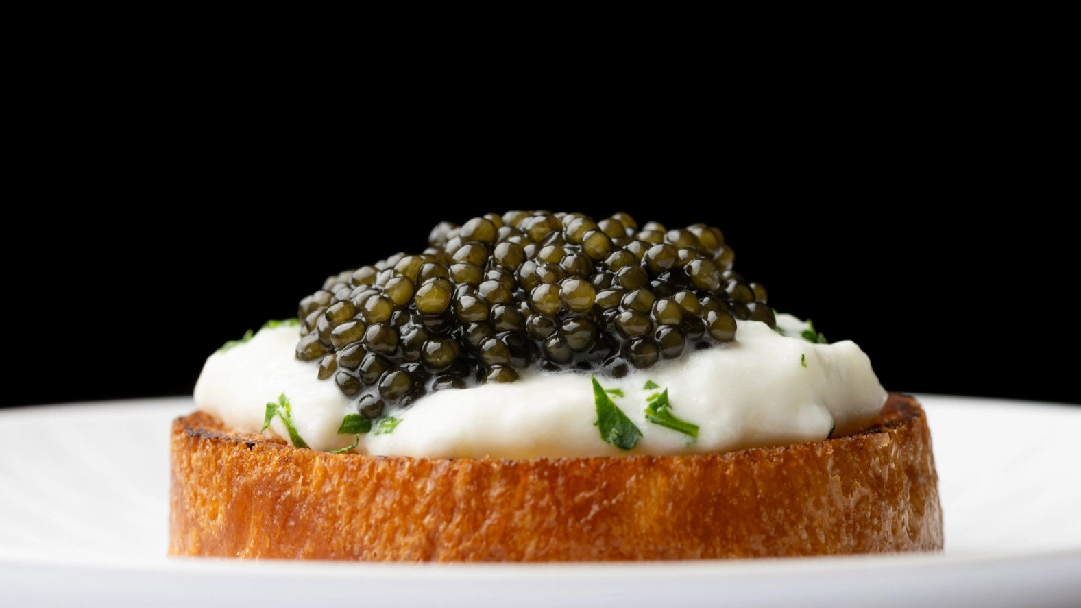 Toasted bread with creamy spread and black caviar, garnished with herbs on a white plate against a black background. Elegant presentation.