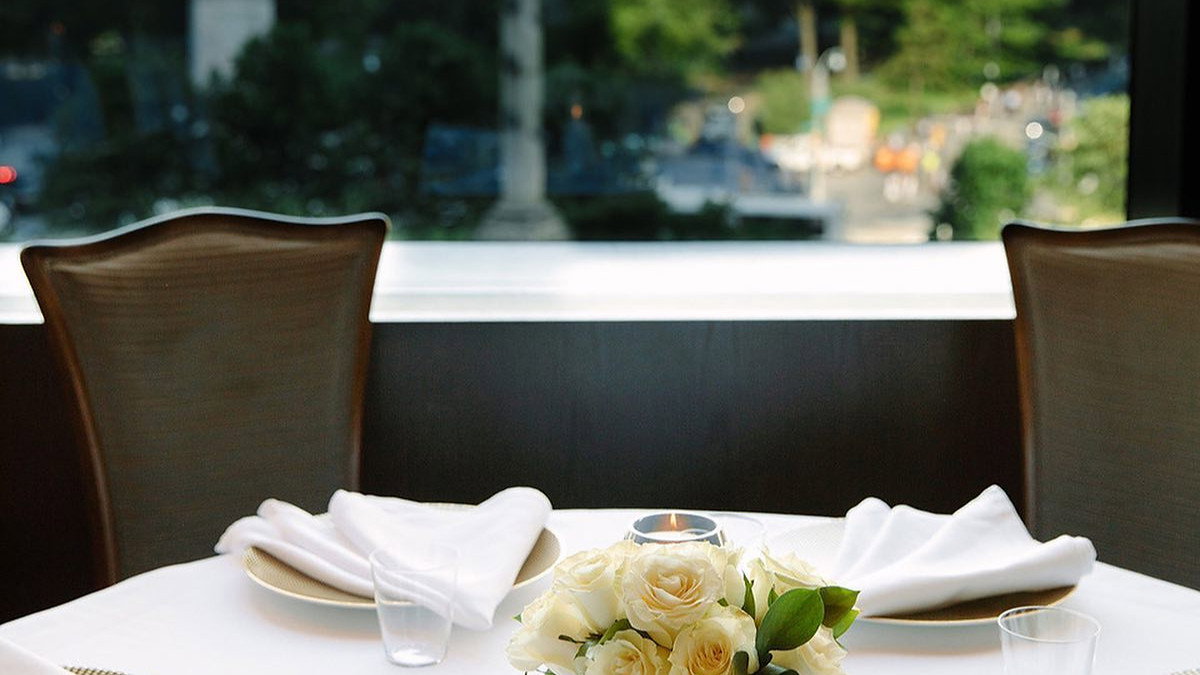 Elegant table setting with white roses and folded napkins, set against a large window overlooking a lush green park and cityscape.