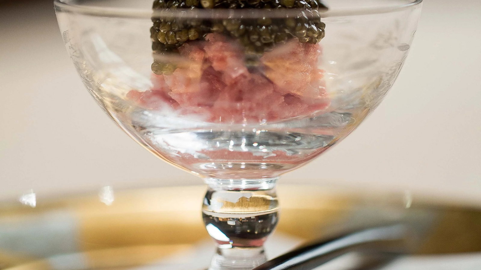 A glass dish with pink tartare and black caviar on top, resting on a gold tray. A spoon is visible in the foreground.