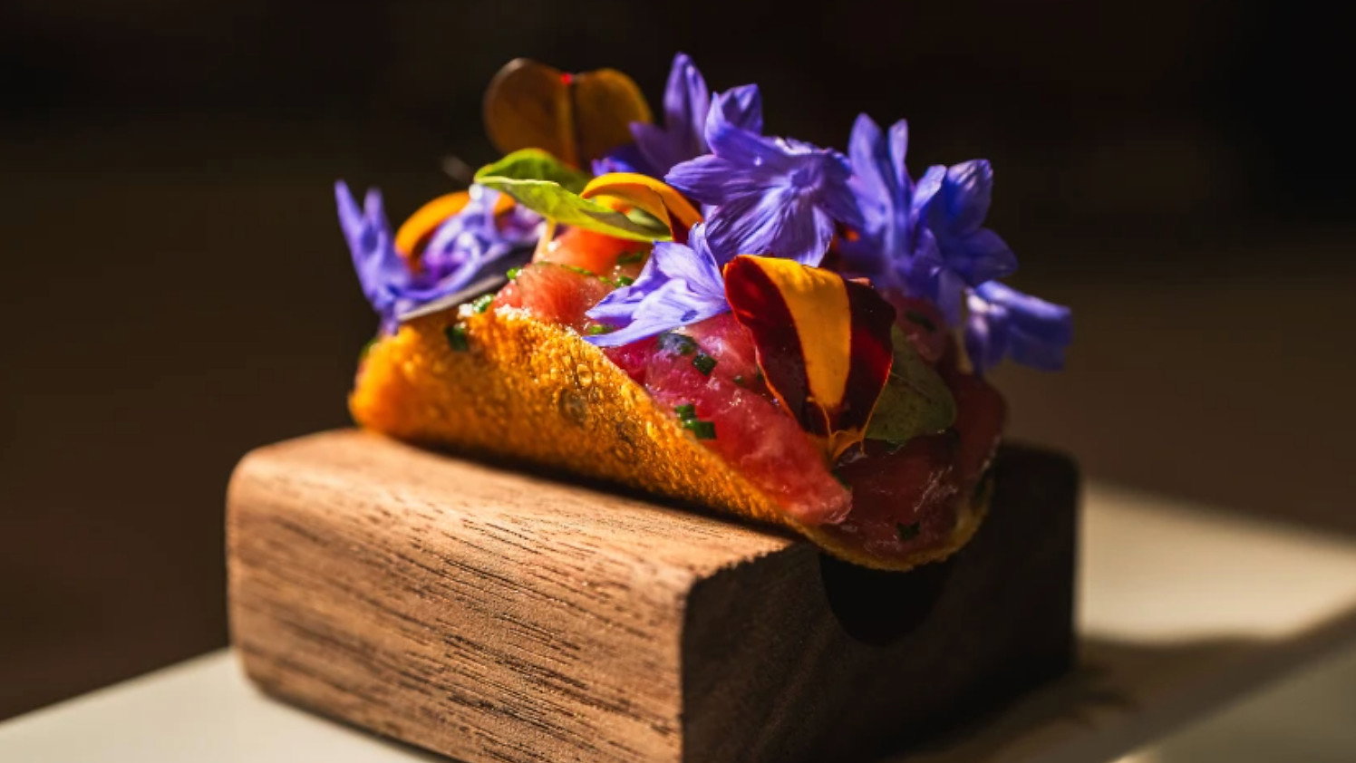 A small taco with vibrant purple and yellow flowers and fresh herbs sits on a wooden block, illuminated against a dark background.