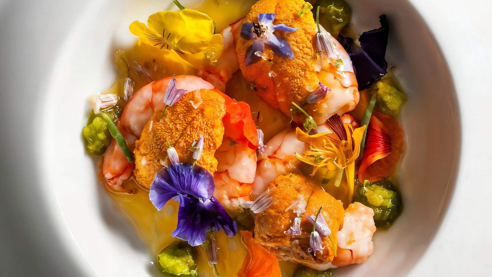 Seafood dish with shrimp and orange roe, garnished with vibrant edible flowers on a white plate. Bright, colorful presentation.