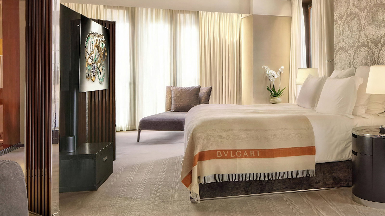 Luxurious hotel room with a large bed, Bvlgari blanket, orchid, and lounge chair. Books and pastries on the table. Soft, warm lighting.