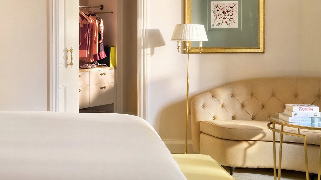 Elegant bedroom with white bed, beige couch, and gold lamp. Open closet with colorful clothes. Framed art on light wall. Cozy ambiance.