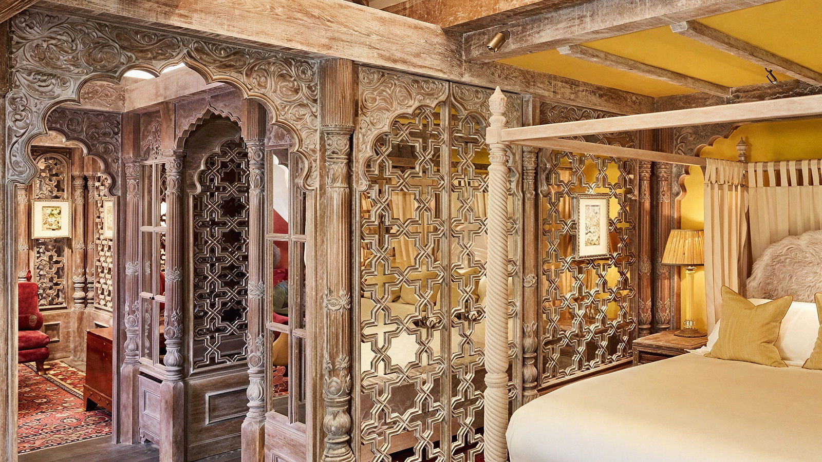 Intricately carved wooden room with a four-poster bed, ornate lattice screens, red rugs, and warm yellow walls. Cozy and elegant ambiance.