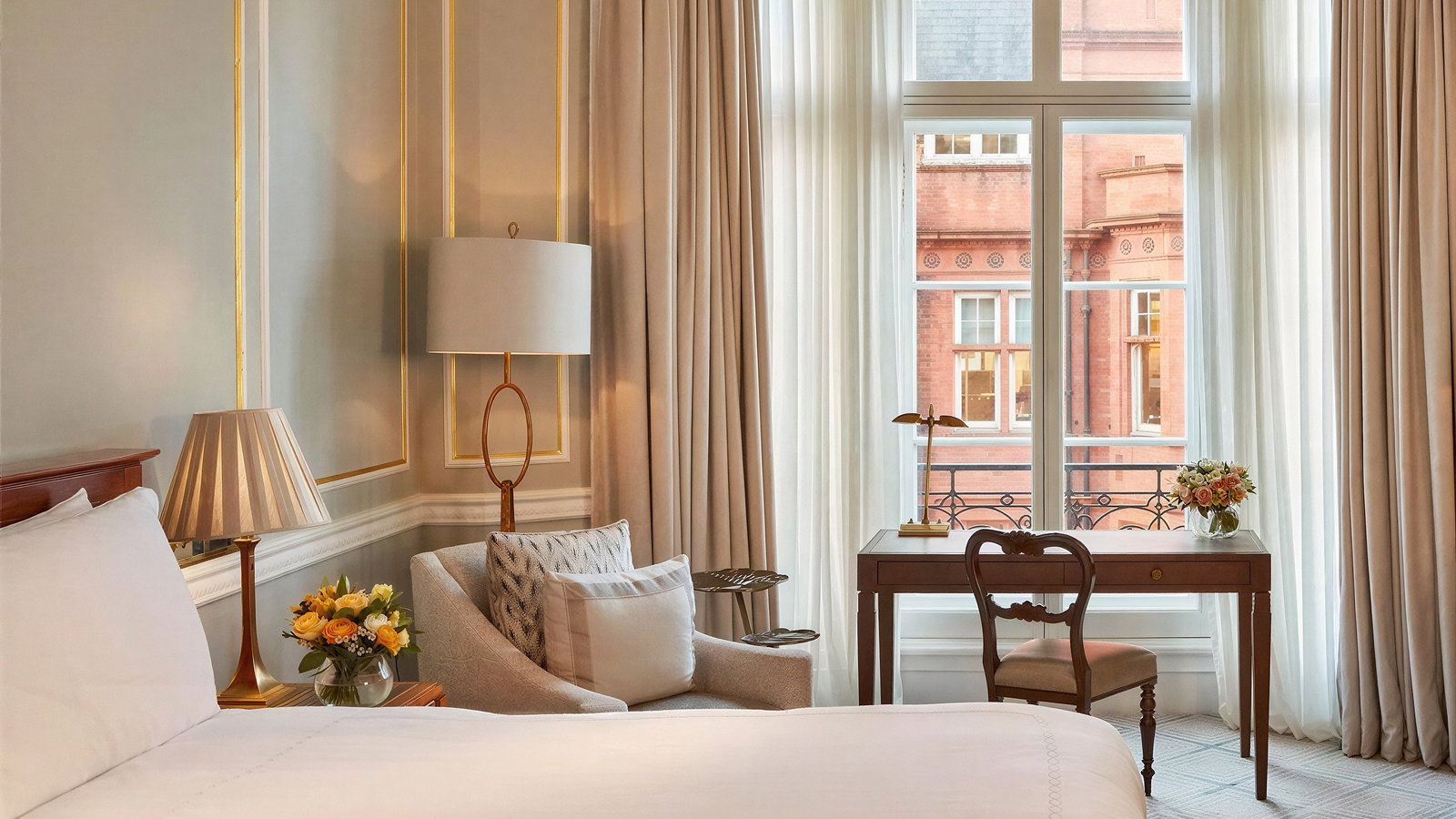 Elegant hotel room with a large bed, soft lighting, and gold accents. A desk by the window reveals a view of red brick buildings outside.