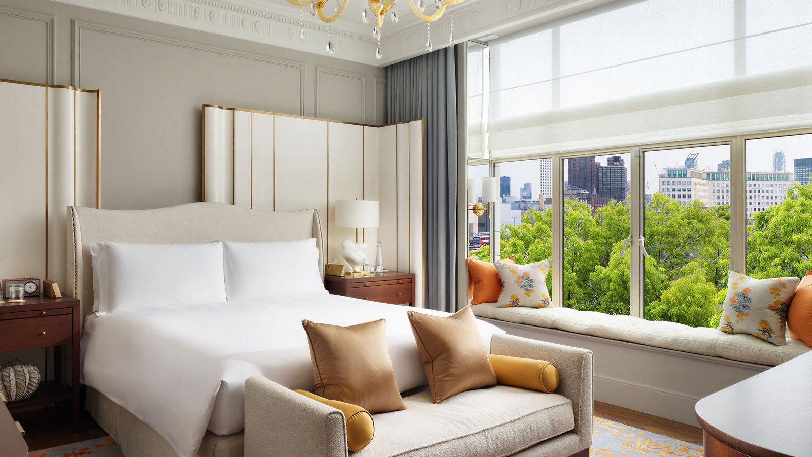 Luxurious hotel room with a king bed, gold chandelier, beige sofa, and city view from large window. Orange accents create a warm ambiance.