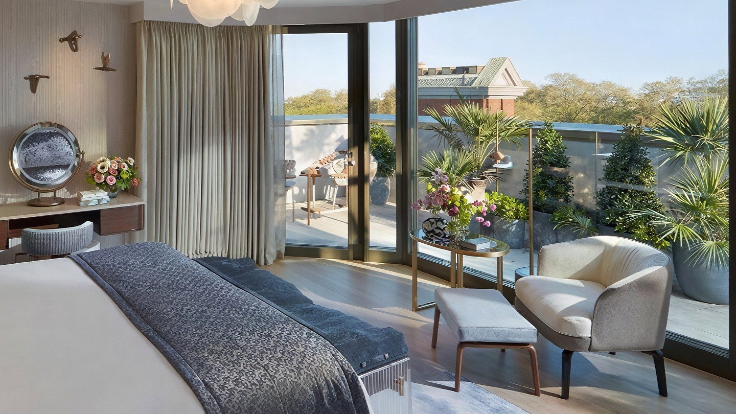 Luxurious bedroom with a large bed, blue patterned throw, and modern decor. Floor-to-ceiling windows reveal a balcony with plants.