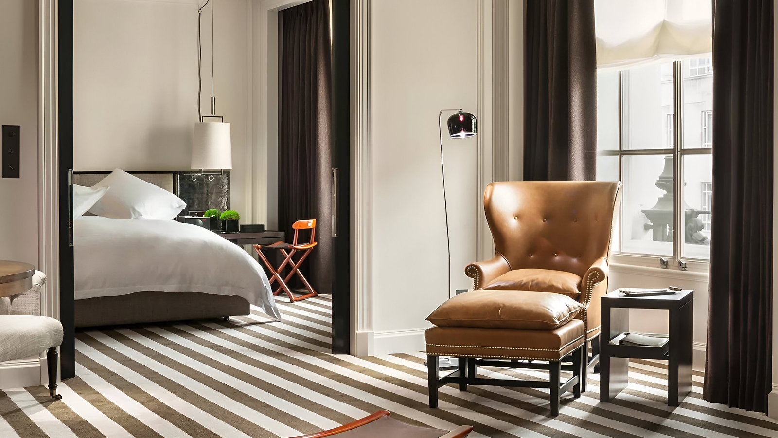 Elegant room with striped carpet, leather chair, and ottoman. A bed is visible through a sliding door. Warm lighting and dark curtains.
